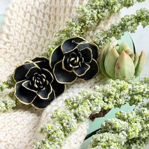 May include: A pair of black and gold hoop earrings with a flower design. The earrings are made of metal and have a black enamel finish. The flowers have a gold outline.