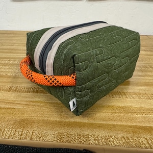 Puede incluir: Una bolsa de tela verde acolchada con cremallera beige y asa de cuerda naranja. La bolsa tiene una etiqueta con el logotipo de un árbol. La cremallera es negra y beige.