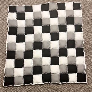 Op de afbeelding: Een zwart-wit geblokte deken met een grijs-wit geblokt patroon. De deken is gemaakt van vierkante stukken stof die aan elkaar zijn genaaid.