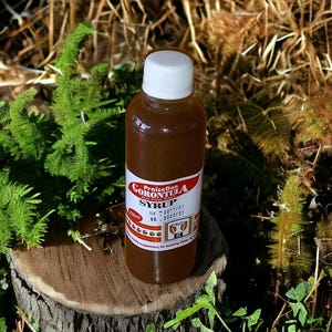 May include: A bottle of brown syrup with a white cap, labelled "PraiseDan Gorontula Syrup". The bottle, containing 250ml, is set against a backdrop of greenery and a wooden surface. The label has red and white details.