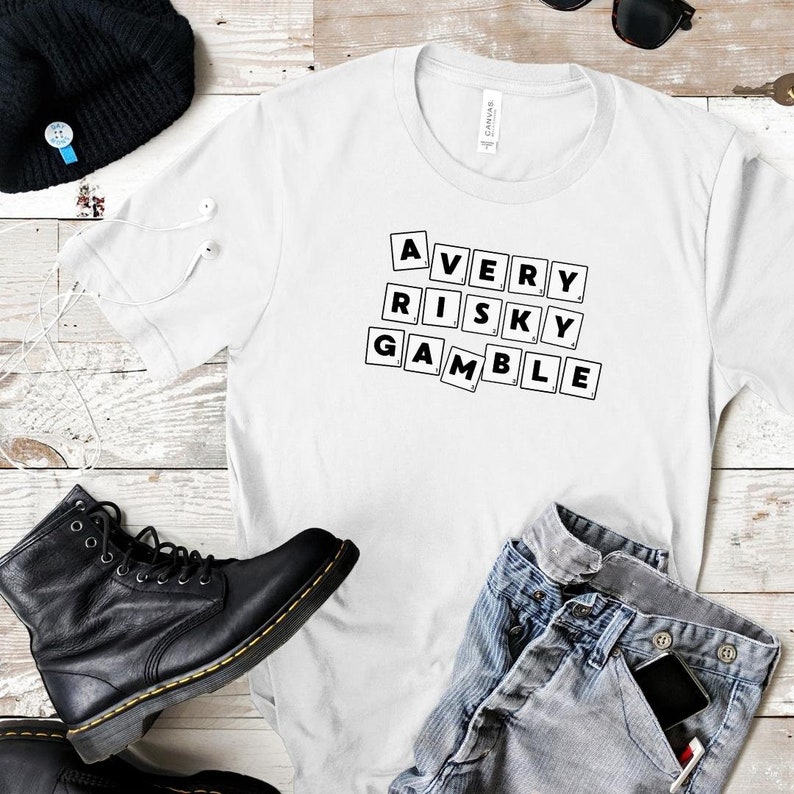 Op de afbeelding: Wit T-shirt met zwarte tekst die "A VERY RISKY GAMBLE" luidt in een bloklettertype. De tekst is trapsgewijs gerangschikt, met elk woord op een aparte regel.