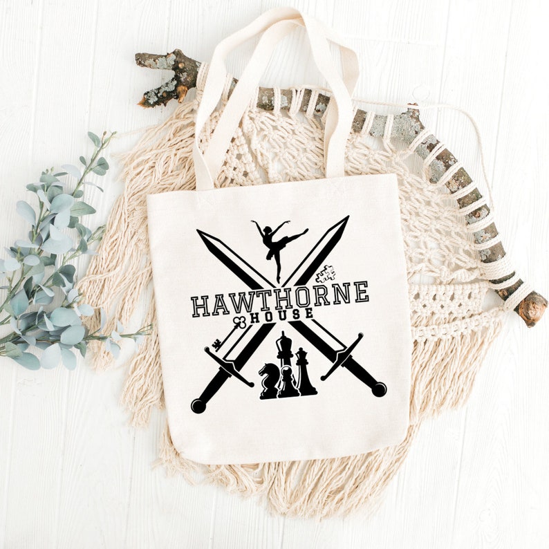 May include: Cream-colored tote bag with a black graphic design. The design includes crossed swords, a ballerina, chess pieces, and the text "HAWTHORNE HOUSE". The bag has two cream-colored handles.