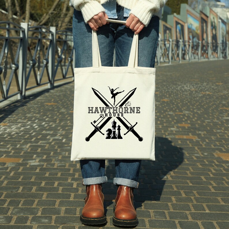 May include: Cream-colored tote bag with a black graphic design. The design features crossed swords, a ballerina, and chess pieces, with the text "HAWTHORNE HOUSE". The bag is held by a person wearing blue jeans and brown boots.
