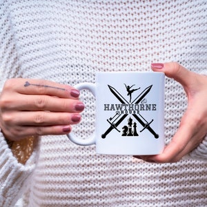 May include: White ceramic mug featuring the text "HAWTHORNE HOUSE" with crossed swords, a ballerina, and chess pieces. The mug is held by a person wearing a white sweater.