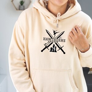 May include: Cream-colored hoodie with a front graphic. The graphic features crossed swords, a ballerina, and chess pieces, with the text "HAWTHORNE HOUSE". The hoodie has a drawstring hood and a front pocket.