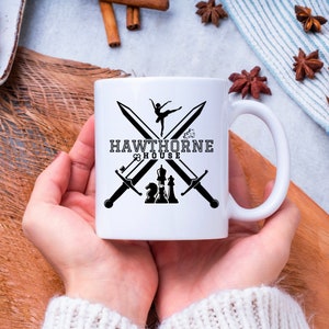 May include: White ceramic mug featuring the text "HAWTHORNE HOUSE" with crossed swords, a ballerina, and chess pieces. The mug is held by a person wearing a white sweater.