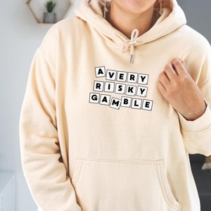 Op de afbeelding: Een cr&egrave;mekleurige hoodie met de woorden "A VERY RISKY GAMBLE" gespeld in zwarte lettertegels.