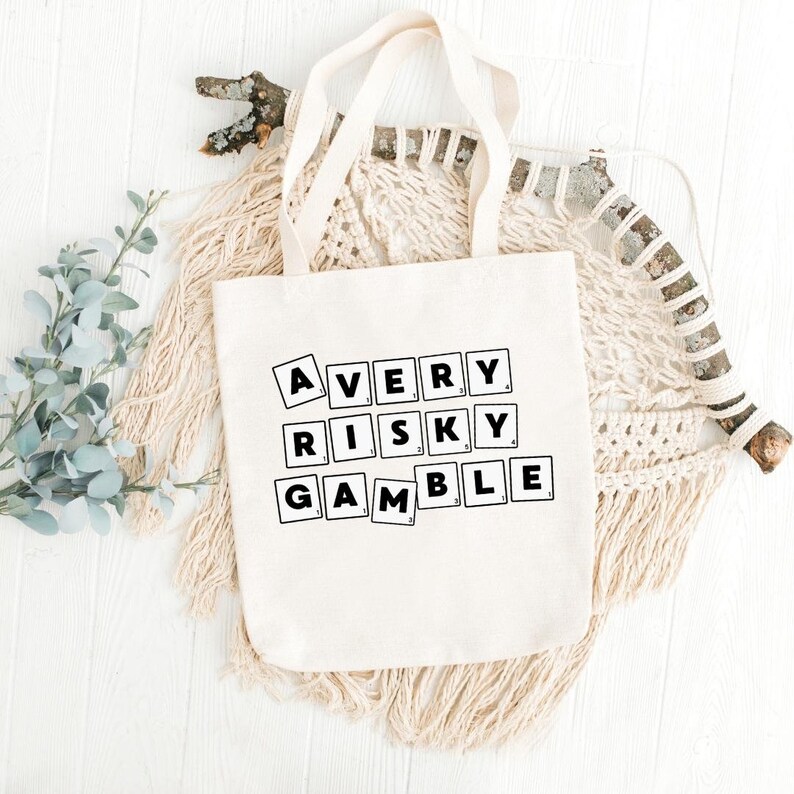 Op de afbeelding: Een witte canvas tote bag met zwarte tekst die "A VERY RISKY GAMBLE" leest in een Scrabble-tegellettertype.