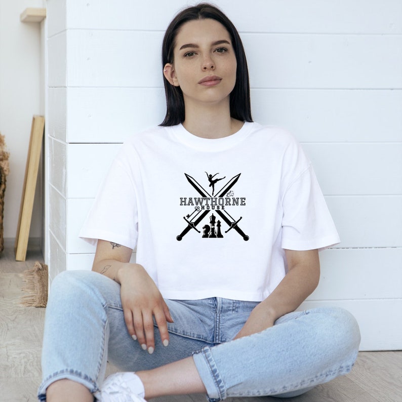 May include: White crop top with a black graphic featuring crossed swords, a ballerina, and chess pieces. The text "Hawthorne House" is centered below the ballerina and swords.