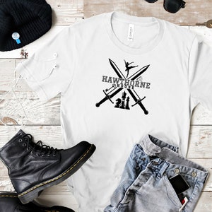 May include: White t-shirt with a black graphic featuring crossed swords, a ballerina, and chess pieces, with the text "HAWTHORNE HOUSE". The shirt is paired with blue jeans, a black boot, and a black beanie.