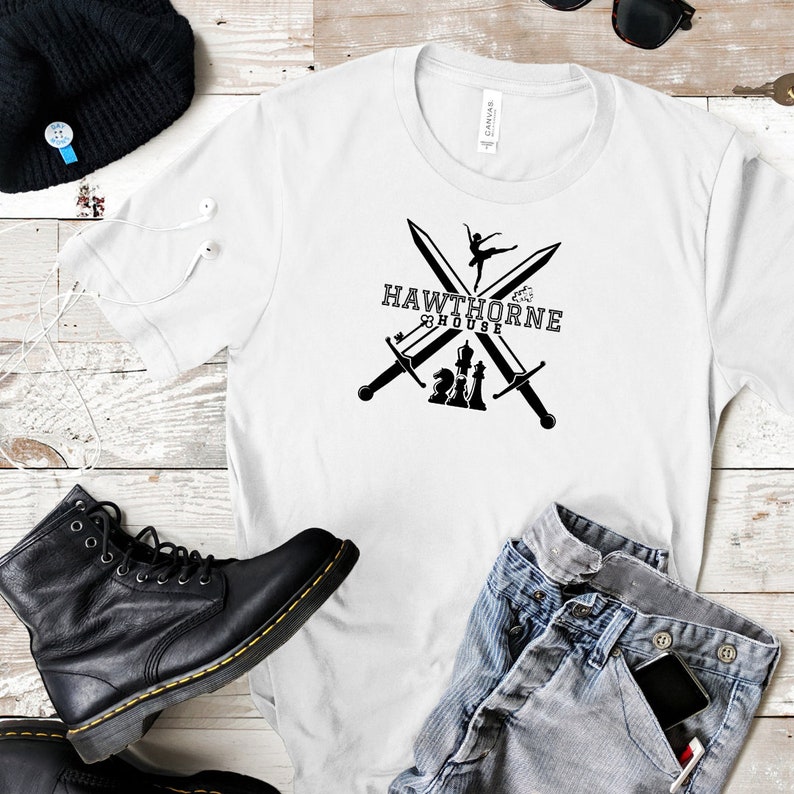 May include: White t-shirt with a black graphic featuring crossed swords, a ballerina, and chess pieces, with the text "HAWTHORNE HOUSE". The shirt is paired with blue jeans, a black boot, and a black beanie.