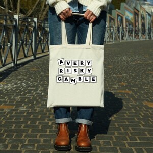 Op de afbeelding: Een witte canvas tote bag met zwarte tekst die "A VERY RISKY GAMBLE" leest in een Scrabble-lettertype.