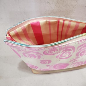 May include: Open, white cosmetic bag with a pink rose pattern. The interior features a pink and white striped lining. The zipper is iridescent. The bag is designed to hold makeup and beauty products.