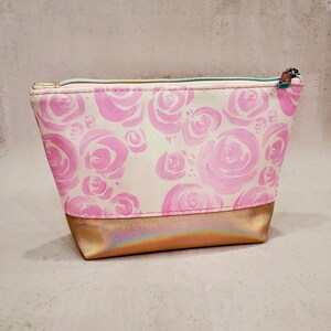 May include: A small, rectangular cosmetic bag with a pink rose pattern on a cream background. The bottom section is a metallic gold color. The bag has a light blue zipper and a small zipper pull.