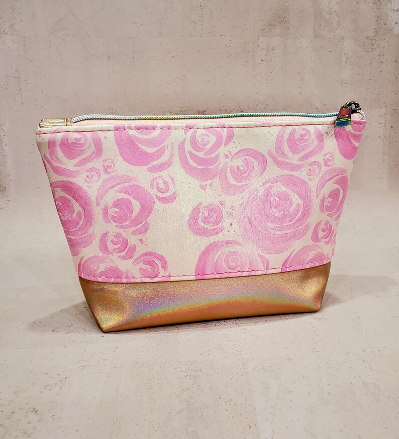 May include: A small, rectangular cosmetic bag with a pink rose pattern on a cream background. The bottom section is a metallic gold color. The bag has a light blue zipper and a small zipper pull.
