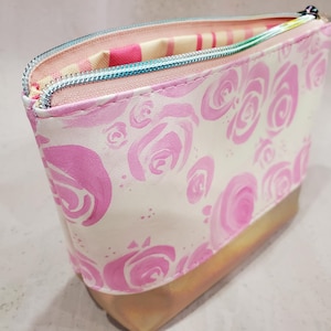 May include: A cosmetic bag with a white background and pink rose pattern. The bag has a gold-colored bottom and a teal zipper. The interior lining is striped pink and white.