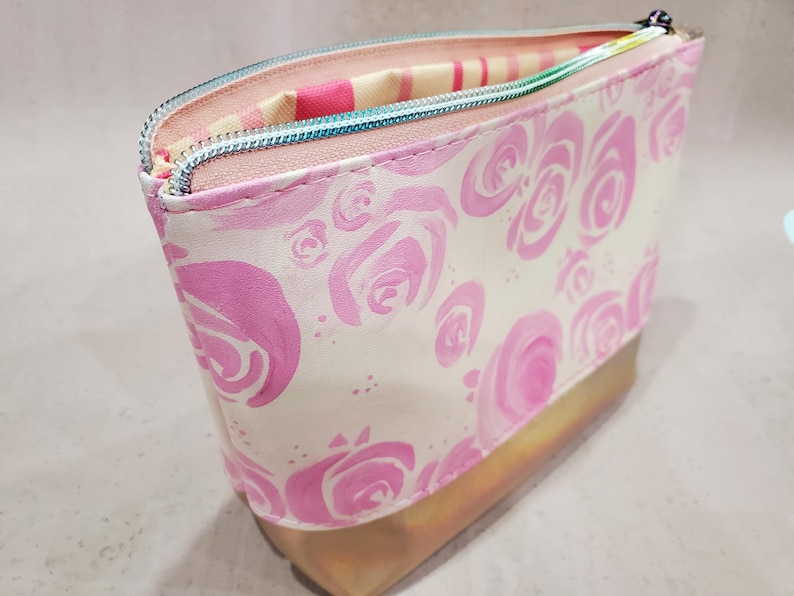 May include: A cosmetic bag with a white background and pink rose pattern. The bag has a gold-colored bottom and a teal zipper. The interior lining is striped pink and white.