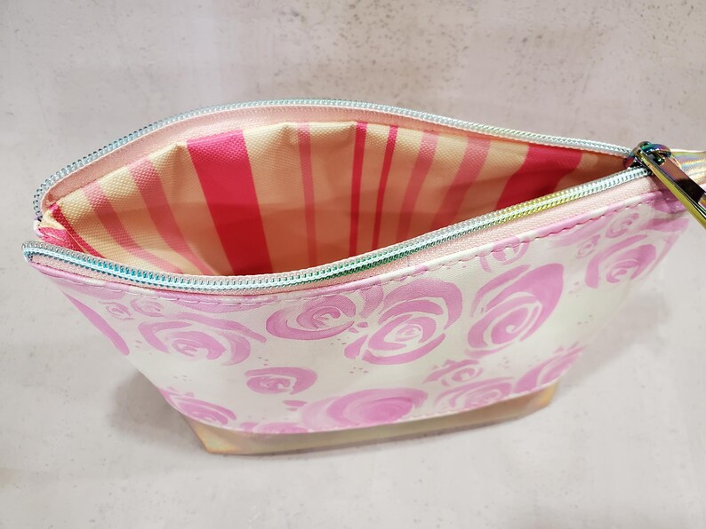 May include: Open, white cosmetic bag with a pink rose pattern. The interior features a pink and white striped lining. The zipper is iridescent. The bag is designed to hold makeup and beauty products.