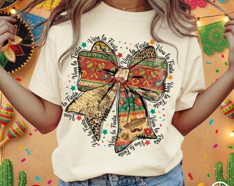 May include: Cream-colored t-shirt featuring a large bow design with a festive theme. The bow incorporates patterns, including maracas, sombreros, and leopard print. The words "Viva la Fiesta" are printed around the bow in a circular pattern.