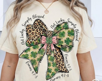 May include: Cream-colored t-shirt featuring a large bow design with leopard print and shamrocks. The bow has a pink center and is surrounded by the text "Not Lucky, Simply Blessed" and "Romans 4:7-8".