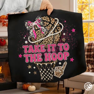 May include: Black blanket with a basketball design. The basketball has a leopard print pattern and is adorned with a pearl necklace and a pink bow. The text "TAKE IT TO THE HOOP" is in pink, with stars and a basketball net graphic.