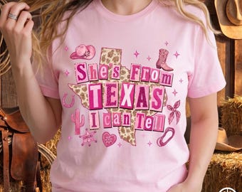 May include: A light pink t-shirt with the text "She's From TEXAS I Can Tell" in pink and white lettering. The design includes a map of Texas with a leopard print pattern, cowboy boots, and other Texas-themed graphics.