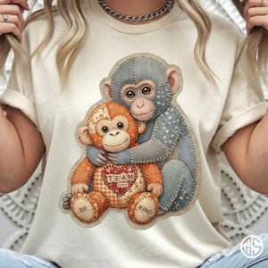 Pode incluir: Camiseta creme com um gráfico de dois macacos de pelúcia costurados abraçando-se. O macaco laranja tem um patch em forma de coração que diz "TEAM PUNCH". O macaco azul abraça o macaco laranja. As palavras "BE" e "KIND" estão no macaco laranja.