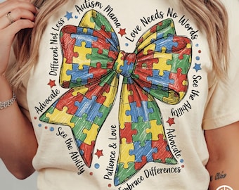 May include: Cream-colored t-shirt featuring a colorful puzzle piece bow design. The bow is made of red, yellow, blue, and green puzzle pieces. Text around the bow includes phrases like "Autism Mama" and "Embrace Differences".