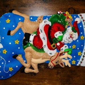 May include: A blue felt Christmas stocking with a white felt cuff. Santa Claus is riding a reindeer with a bag of presents. The stocking is decorated with white snowflakes, yellow stars, and green sequins.