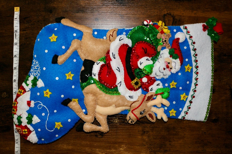 May include: A blue felt Christmas stocking with a white felt cuff. Santa Claus is riding a reindeer with a bag of presents. The stocking is decorated with white snowflakes, yellow stars, and green sequins.
