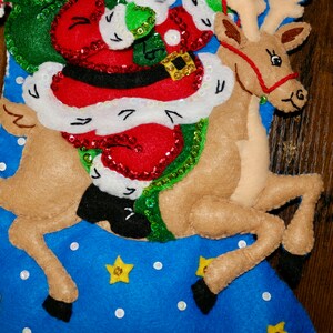 May include: A Christmas stocking with a blue background and white polka dots. Santa Claus is riding a reindeer with a red harness. The reindeer is brown with black hooves. There are yellow stars on the stocking.