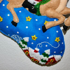 May include: A blue felt stocking with a reindeer applique. The stocking is decorated with white snowflakes, white dots, and yellow stars. The reindeer is brown with a green and red scarf.