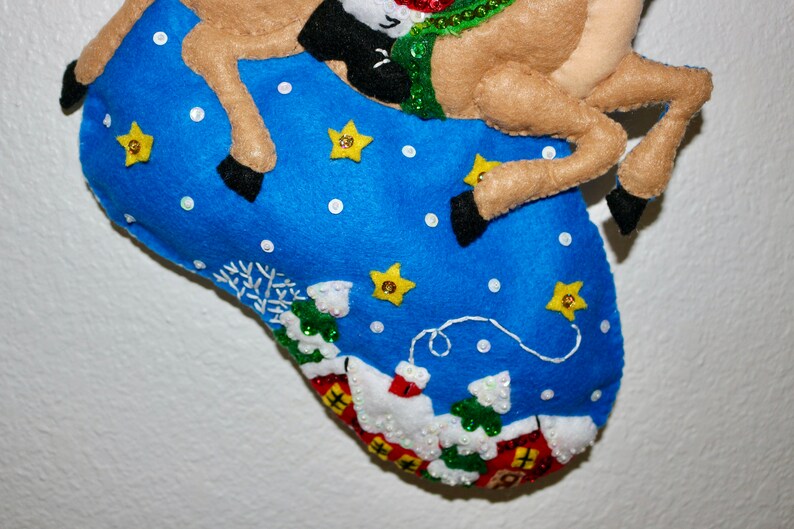 May include: A blue felt stocking with a reindeer applique. The stocking is decorated with white snowflakes, white dots, and yellow stars. The reindeer is brown with a green and red scarf.