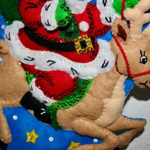 May include: A felt Christmas stocking with a reindeer and Santa Claus design. The reindeer is brown and has a red nose. Santa Claus is wearing a red suit with white trim and a green hat. The stocking is blue with white stars and sequins.