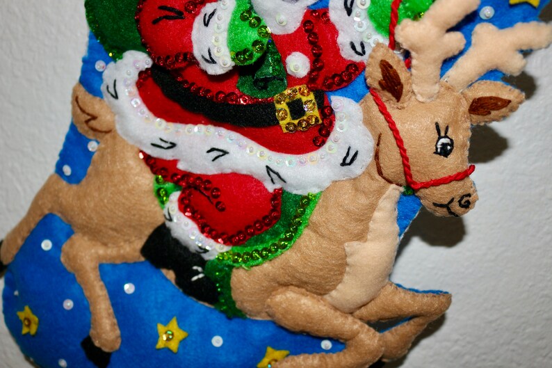 May include: A felt Christmas stocking with a reindeer and Santa Claus design. The reindeer is brown and has a red nose. Santa Claus is wearing a red suit with white trim and a green hat. The stocking is blue with white stars and sequins.