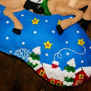 May include: A felt Christmas stocking with a reindeer and a winter village scene. The stocking is blue with white snowflakes and yellow stars. The reindeer is brown with black hooves and a green and white scarf. The village scene includes small white houses with red roofs and green trees.