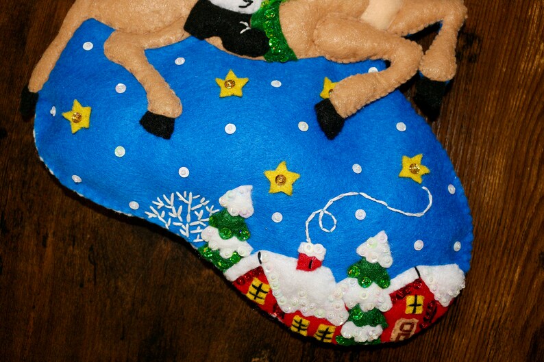 May include: A felt Christmas stocking with a reindeer and a winter village scene. The stocking is blue with white snowflakes and yellow stars. The reindeer is brown with black hooves and a green and white scarf. The village scene includes small white houses with red roofs and green trees.