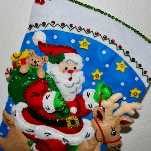 May include: A blue and white Christmas stocking with a Santa Claus, reindeer, and teddy bear design. The stocking is decorated with sequins, beads, and stars.