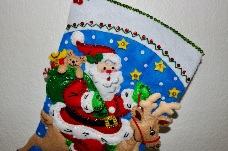 May include: A blue and white Christmas stocking with a Santa Claus, reindeer, and teddy bear design. The stocking is decorated with sequins, beads, and stars.