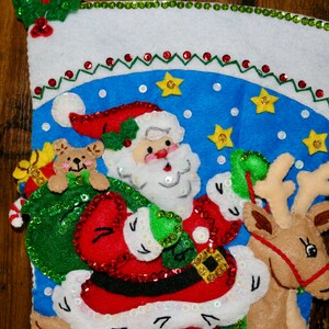 May include: A Christmas stocking with a blue background and a Santa Claus figure holding a bag of presents and riding a reindeer. The stocking is decorated with sequins and stars.