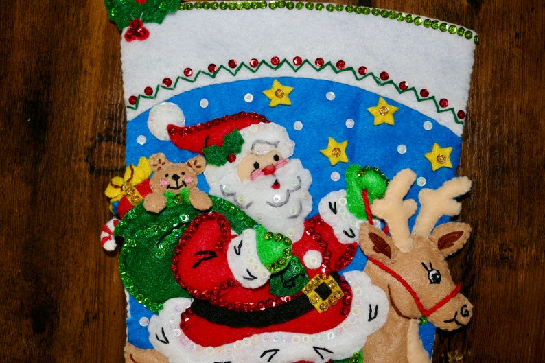 May include: A Christmas stocking with a blue background and a Santa Claus figure holding a bag of presents and riding a reindeer. The stocking is decorated with sequins and stars.