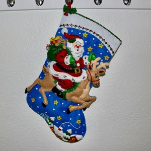 May include: A blue Christmas stocking with a Santa Claus and reindeer design. Santa is holding a bag of gifts and riding the reindeer. The stocking is decorated with stars and sequins.