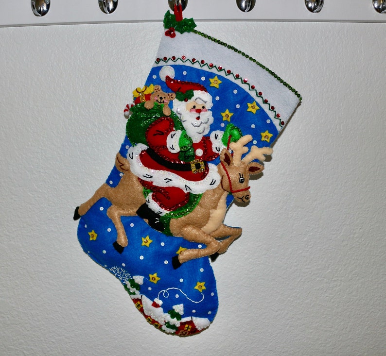 May include: A blue Christmas stocking with a Santa Claus and reindeer design. Santa is holding a bag of gifts and riding the reindeer. The stocking is decorated with stars and sequins.