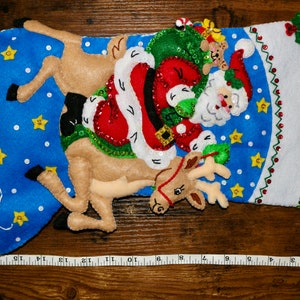 May include: A blue felt Christmas stocking with a Santa Claus and reindeer design. Santa is holding a green bag with a teddy bear peeking out. The reindeer is brown and has a red nose. The stocking is decorated with white snowflakes, yellow stars, and green sequins.