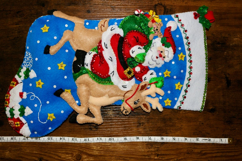 May include: A blue felt Christmas stocking with a Santa Claus and reindeer design. Santa is holding a green bag with a teddy bear peeking out. The reindeer is brown and has a red nose. The stocking is decorated with white snowflakes, yellow stars, and green sequins.