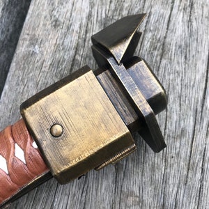 Puede incluir: Primer plano de una empu&ntilde;adura de espada de metal de color dorado con una vaina de cuero marr&oacute;n. La empu&ntilde;adura tiene una forma cuadrada con un peque&ntilde;o bot&oacute;n redondo en el lateral. La vaina tiene una franja blanca que recorre el centro.