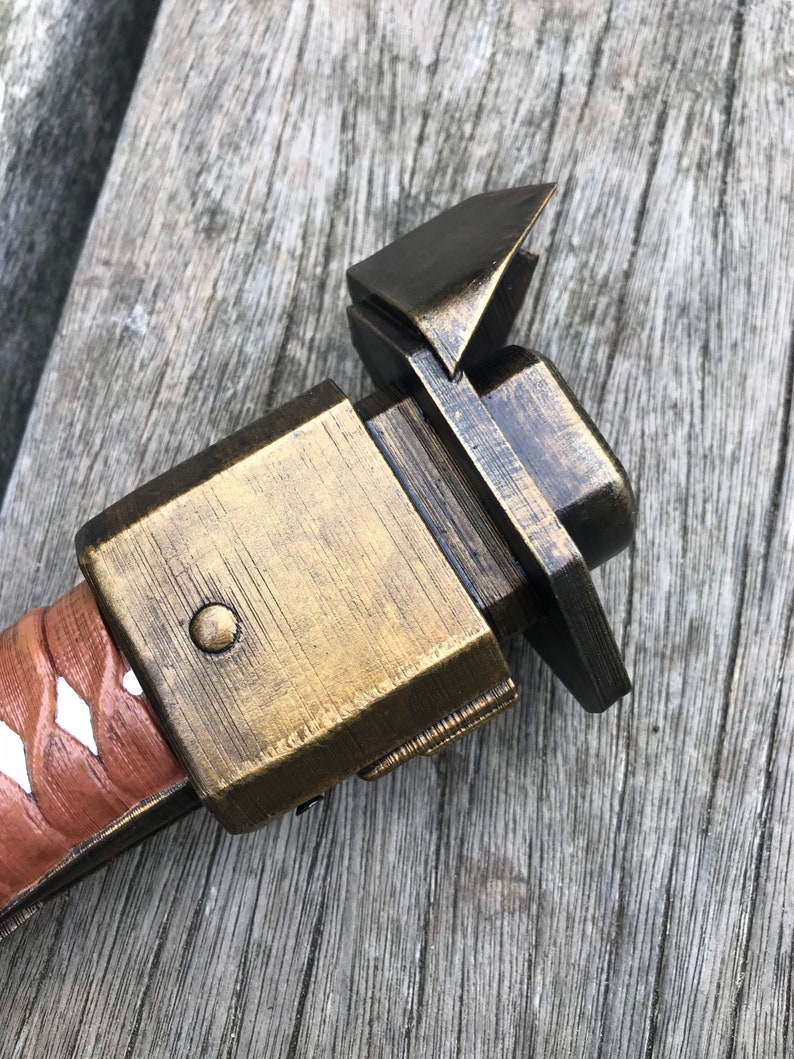 Puede incluir: Primer plano de una empu&ntilde;adura de espada de metal de color dorado con una vaina de cuero marr&oacute;n. La empu&ntilde;adura tiene una forma cuadrada con un peque&ntilde;o bot&oacute;n redondo en el lateral. La vaina tiene una franja blanca que recorre el centro.