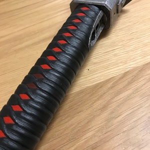 Puede incluir: Primer plano de un mango de katana negro y rojo con una empu&ntilde;adura de metal. El mango est&aacute; envuelto en cuero negro con patrones de diamantes rojos.
