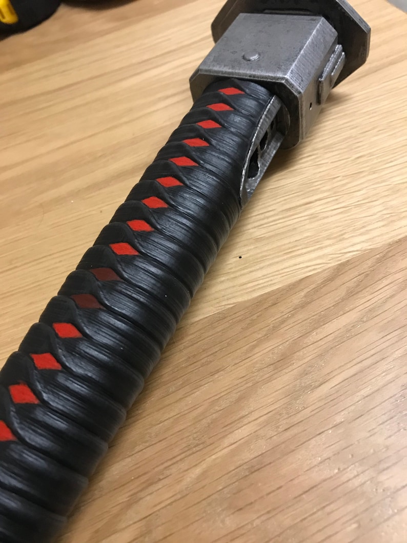 Puede incluir: Primer plano de un mango de katana negro y rojo con una empu&ntilde;adura de metal. El mango est&aacute; envuelto en cuero negro con patrones de diamantes rojos.