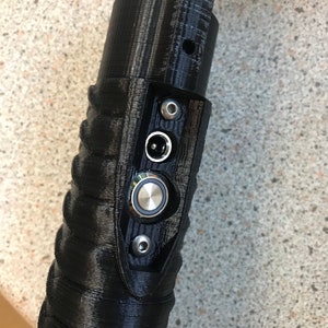 Puede incluir: Primer plano de una empu&ntilde;adura de sable l&aacute;ser negra con un bot&oacute;n plateado y un conector de cable de alimentaci&oacute;n negro.